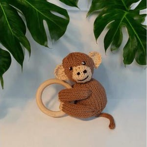 May include: A brown knitted monkey toy with a cream-colored face, ears, and hands, featuring black embroidered eyes and a mouth. The monkey is holding a smooth, round, light-colored wooden ring in its arms and has a long, thin, knitted tail.