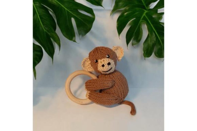 May include: A brown knitted monkey toy with a cream-colored face, ears, and hands, featuring black embroidered eyes and a mouth. The monkey is holding a smooth, round, light-colored wooden ring in its arms and has a long, thin, knitted tail.