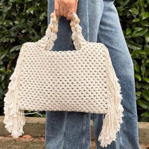 May include: A cream-colored macrame handbag featuring a woven diamond pattern, long fringe detailing on both sides, and two handles accented with small, round, light-colored wooden beads integrated into the knotted cord structure.