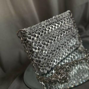 Könnte beinhalten: Eine rechteckige Handtasche aus miteinander verbundenen silbernen Getränkedosen-Laschen, die mit metallisch grauer Kordel verwebt sind. Die Tasche hat einen strukturierten Klappverschluss und einen silberfarbenen Metallkettenriemen an den Seiten.