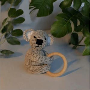 May include: A beige knitted koala toy featuring white inner ears, a black embroidered nose and mouth, and a smooth, circular light-colored wooden ring held by the koala's arms. The toy is crafted with a textured knit pattern throughout its body.