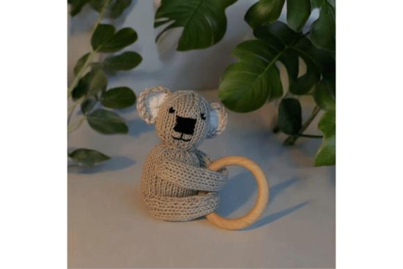 May include: A beige knitted koala toy featuring white inner ears, a black embroidered nose and mouth, and a smooth, circular light-colored wooden ring held by the koala's arms. The toy is crafted with a textured knit pattern throughout its body.
