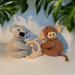 May include: A light gray knitted koala and a brown knitted monkey, each holding a smooth, round wooden ring. The koala has a black stitched nose and ears, while the monkey features a lighter brown face, stitched facial details, and a long tail.