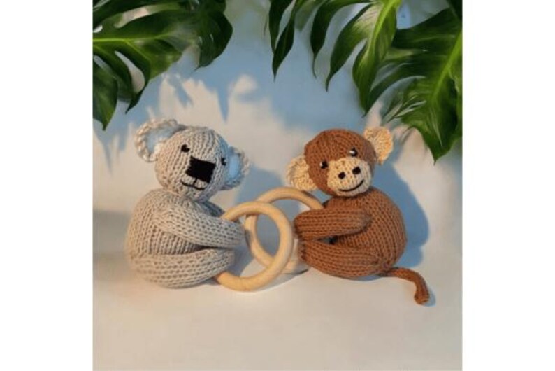 May include: A light gray knitted koala and a brown knitted monkey, each holding a smooth, round wooden ring. The koala has a black stitched nose and ears, while the monkey features a lighter brown face, stitched facial details, and a long tail.