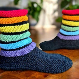 Puede incluir: Un par de calcetines tejidos a mano con el pie de color negro sólido y un puño acanalado multicolor. El puño consta de bandas horizontales apiladas en rojo, naranja, amarillo, verde claro, azul claro y morado, separadas por finas rayas negras.