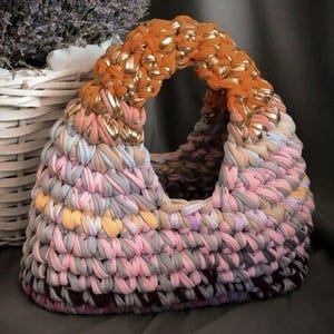 May include: Handmade crochet handbag in shades of pink, gray, yellow, and brown. The handle is orange with gold accents. The bag is next to a white wicker basket with dried lavender.