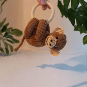 May include: A brown knitted monkey toy with a cream-colored face and ears, hanging from a smooth, round, light-colored wooden ring. The monkey has a long tail and is crafted with visible knit stitches, featuring dark eyes and a stitched mouth.
