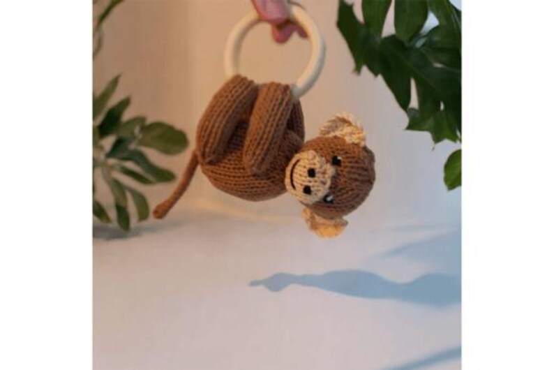May include: A brown knitted monkey toy with a cream-colored face and ears, hanging from a smooth, round, light-colored wooden ring. The monkey has a long tail and is crafted with visible knit stitches, featuring dark eyes and a stitched mouth.