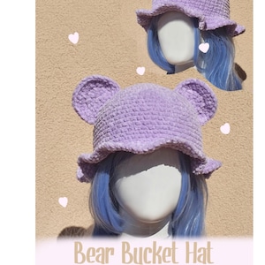 Puede incluir: Un sombrero de cubo de oso de ganchillo de color lila con orejas de oso y un borde con volantes. El sombrero se muestra en una cabeza de maniquí con pelo azul. El texto "Bear Bucket Hat Crochet Pattern" es visible.
