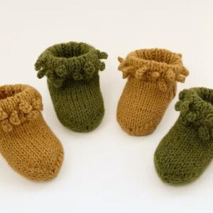 May include: Four hand-knitted booties arranged in a group. Two booties are olive green and two are mustard yellow. Each bootie features a ribbed cuff with a decorative bobble texture around the top edge and a smooth knit foot section.