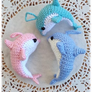 May include: Three handmade crochet dolphins in shades of blue and pink, with white bellies and black eyes. The dolphins are arranged on a white, lacy surface, showcasing their detailed craftsmanship. A charming, whimsical ocean-themed craft.