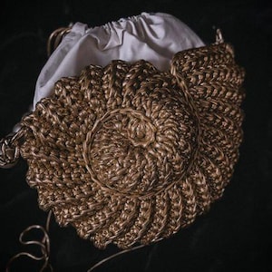May include: A metallic gold crocheted handbag shaped like a seashell with a spiral pattern. The bag features a white fabric drawstring interior lining and a thin gold cord strap, showcasing a textured, woven construction throughout the exterior.