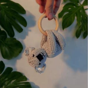 May include: A beige knitted koala toy attached to a smooth, round, light-colored wooden ring. The koala features a textured knit body, embroidered black eyes, and a black nose, with small ears and limbs wrapped around the wooden ring structure.