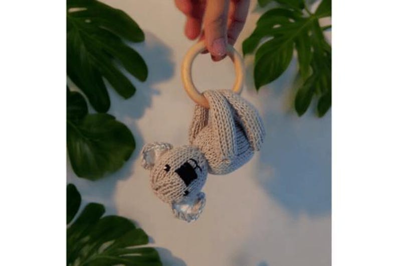 May include: A beige knitted koala toy attached to a smooth, round, light-colored wooden ring. The koala features a textured knit body, embroidered black eyes, and a black nose, with small ears and limbs wrapped around the wooden ring structure.