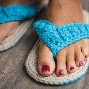 Könnte beinhalten: Ein Paar handgehäkelte Flip-Flops mit leuchtend türkisfarbenen Garnriemen in strukturiertem Muster und einer Sohle aus beigem Garn. Die Basis der Schuhe ist mit einem passenden türkisfarbenen Rand entlang der Außenkante versehen.