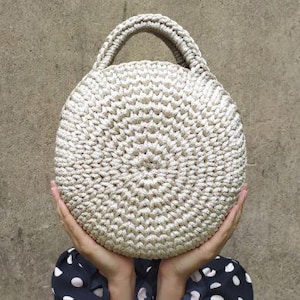 May include: A cream-colored, round crochet handbag with a sturdy handle. The bag's texture is visible, showcasing the detailed stitching. The bag is held up by a person. The background is a neutral, textured wall.