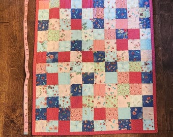 Op de afbeelding: Een kleine, vierkante patchworkquilt met een roze rand. De quilt is gemaakt van verschillende vierkante stoffen met bloemenpatronen en effen kleuren in roze, blauw en groen. De quilt meet ongeveer 69 cm lang.