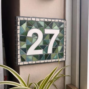 May include: A rectangular mosaic house number plaque displaying the number 27 in white. The mosaic features green, blue, and white tiles, framed by a white border. A decorative wall sconce is positioned above the plaque.