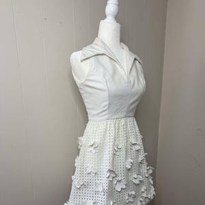 May include: A white sleeveless dress with a collared neckline and a skirt with a floral design. The skirt is made of a white fabric with a textured pattern.