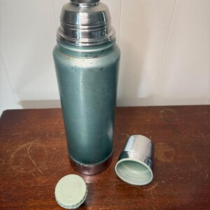 Puede incluir: Un termo verde vintage con una tapa plateada y una tapa verde separada. El termo est&aacute; sobre una superficie de madera.