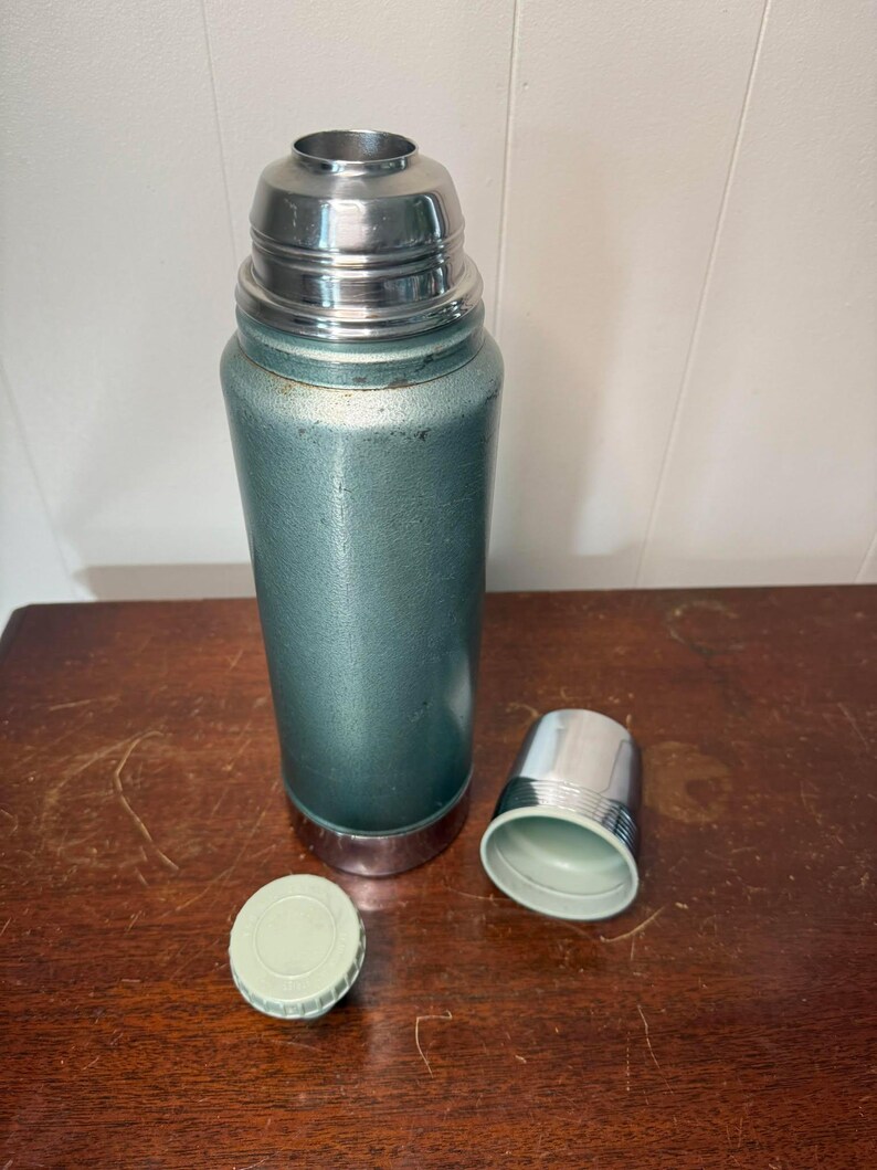 Puede incluir: Un termo verde vintage con una tapa plateada y una tapa verde separada. El termo est&aacute; sobre una superficie de madera.