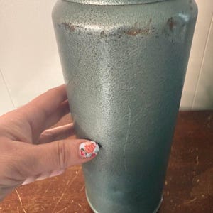 Puede incluir: Un termo vintage de metal de color verde azulado con una tapa plateada y una base marr&oacute;n. El termo tiene una superficie texturizada y muestra signos de desgaste.