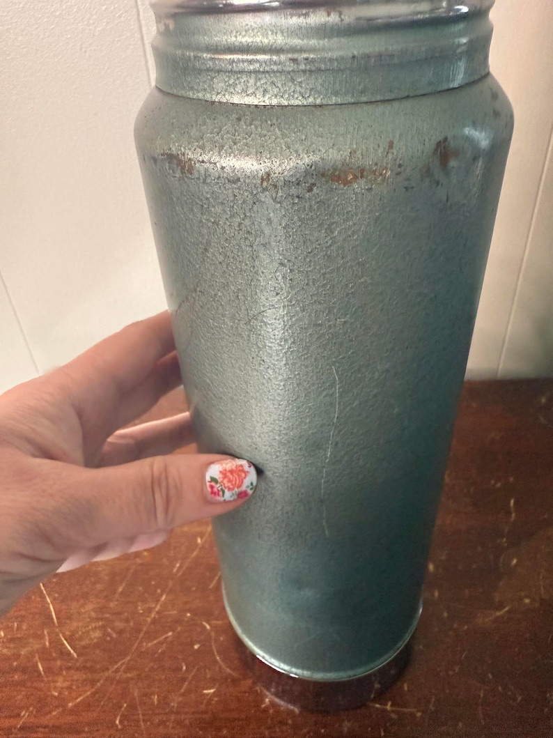 Puede incluir: Un termo vintage de metal de color verde azulado con una tapa plateada y una base marr&oacute;n. El termo tiene una superficie texturizada y muestra signos de desgaste.