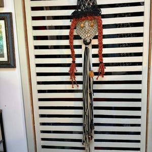 May include: A macrame wall hanging featuring a black pointed hat, a textured beige face with round wooden eyes, and two long braided rust-colored yarn pigtails. Below the face, a long fringe of black, white, and beige yarn hangs down with wooden beads.