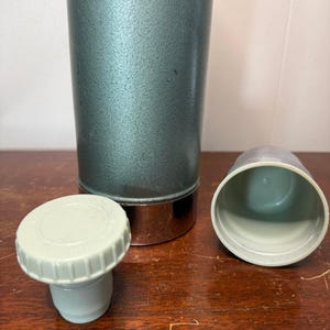 Puede incluir: Un termo vintage de metal verde con fondo cromado y tapa y tap&oacute;n de pl&aacute;stico blanco.