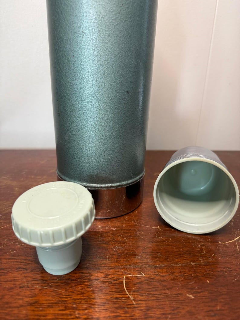 Puede incluir: Un termo vintage de metal verde con fondo cromado y tapa y tap&oacute;n de pl&aacute;stico blanco.