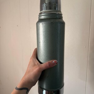 Puede incluir: Un termo vintage plateado con tapa y base cromadas. El termo tiene una superficie texturizada y est&aacute; siendo sostenido por una mano.