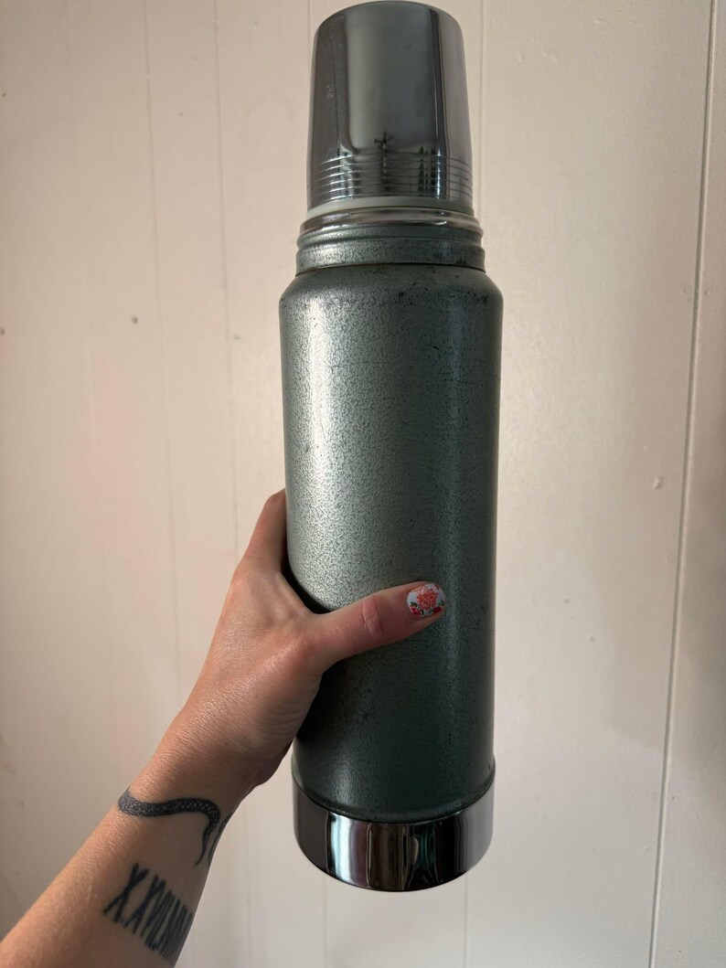 Puede incluir: Un termo vintage plateado con tapa y base cromadas. El termo tiene una superficie texturizada y est&aacute; siendo sostenido por una mano.