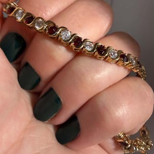 May include: A gold-tone bracelet featuring an alternating pattern of round clear stones and round deep red stones set within a continuous S-shaped metal link design. The bracelet has a secure clasp mechanism visible at the end of the chain.