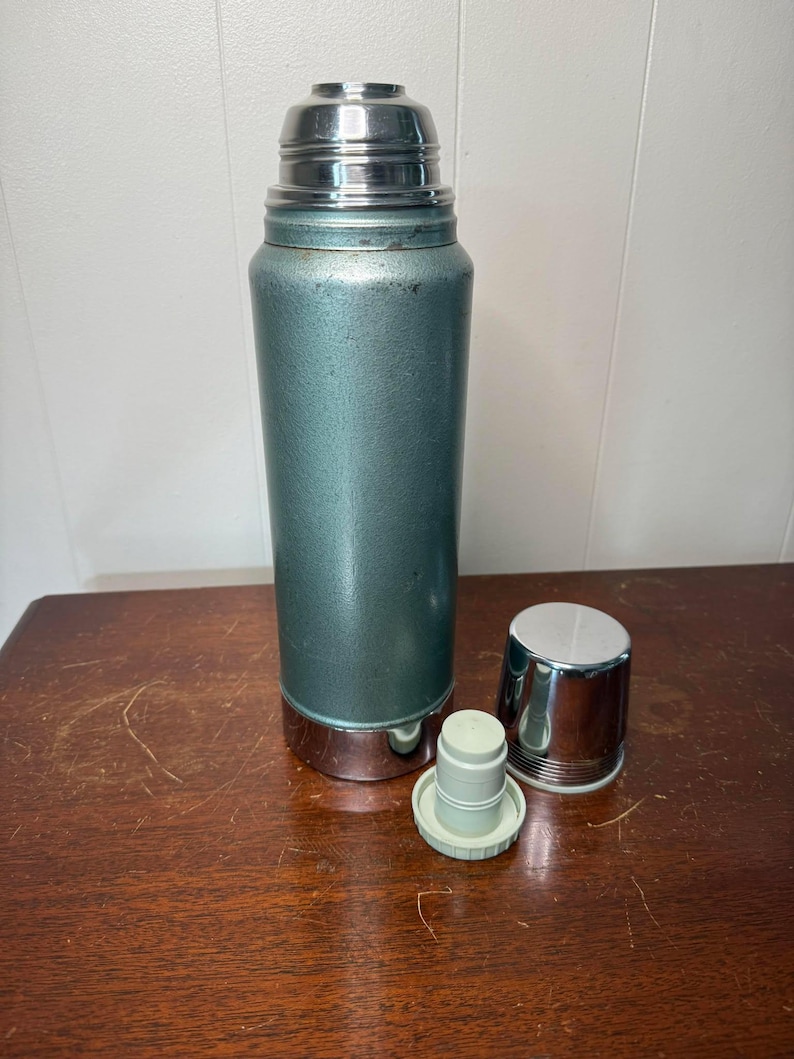 Puede incluir: Un termo vintage azul y plateado con una tapa extra&iacute;ble y un tap&oacute;n separado. El termo tiene una superficie texturizada y una banda plateada alrededor de la parte inferior.