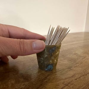 May include: A small, cylindrical brass container with an etched floral pattern, filled with a cluster of light-colored wooden toothpicks. The metal surface shows a weathered patina with dark, oxidized areas throughout the engraved design.