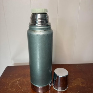 Puede incluir: Un termo vintage de metal verde con tapa plateada y tap&oacute;n plateado. El termo est&aacute; sobre una superficie de madera.