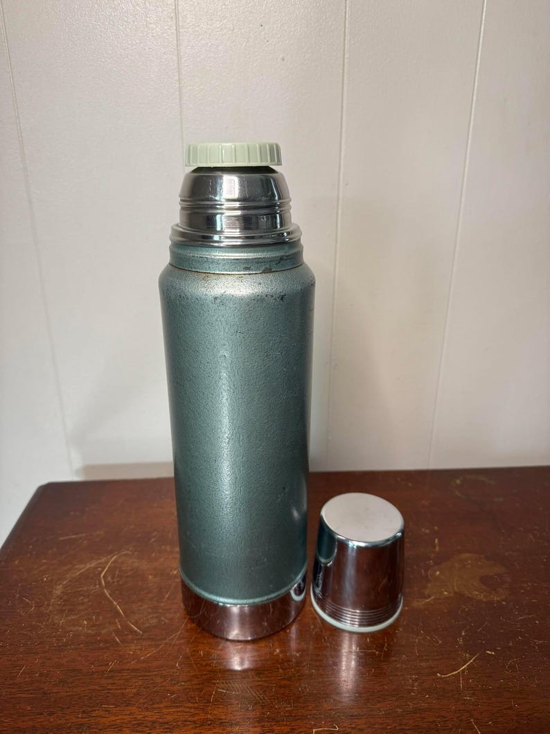 Puede incluir: Un termo vintage de metal verde con tapa plateada y tap&oacute;n plateado. El termo est&aacute; sobre una superficie de madera.