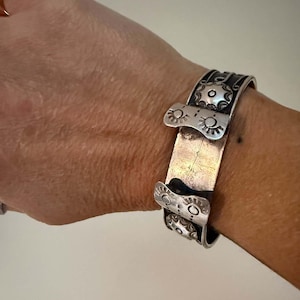 May include: A silver-toned cuff bracelet featuring a wide, flat central band flanked by two decorative plates. Each plate displays embossed sunburst motifs and circular patterns with dark, recessed detailing against a polished metal surface.