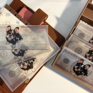 May include: Clear plastic money envelopes with a French Bulldog design, featuring a $100 bill background. The envelopes are arranged with a brown leather wallet and a wooden organizer.