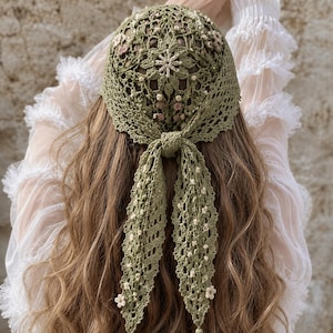 May include: A sage green crochet head covering with a floral design and trailing ties. The head covering is adorned with small white and pink floral accents. The intricate openwork design is visible, showcasing the craftsmanship of the handmade piece.