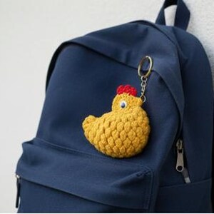 May include: A yellow crochet chicken keychain with a red comb and blue eyes is attached to a navy blue backpack. The keychain is made of textured yarn and has a metal keyring.