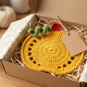 May include: A handmade yellow crochet pumpkin coaster with green vine and red accents, inside a cardboard box with decorative paper and a blank tag. The coaster is a circular shape with decorative holes.