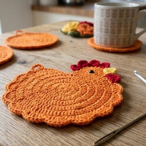 Può includere: Sottobicchieri all'uncinetto arancioni fatti a mano e un sottopentola a forma di gallina su un tavolo di legno. Una tazza poggia su un sottobicchiere, con filato e uncinetti nelle vicinanze. Il sottopentola ha dettagli rossi, gialli e neri.