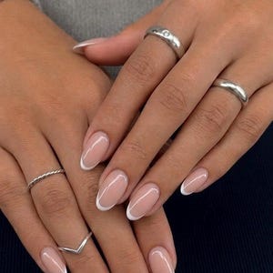 Peut inclure: Un ensemble de quatre bagues argentées portées sur des doigts aux ongles en amande arborant une manucure française classique avec une base nude et des pointes blanches. Les bagues incluent un anneau simple avec une petite pierre, un anneau lisse, un anneau torsadé et un anneau en V.