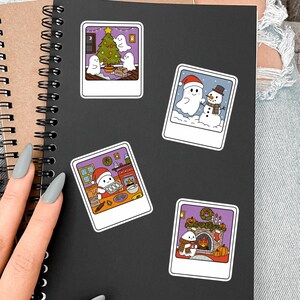 May include: A black spiral-bound notebook decorated with four polaroid-style stickers featuring cartoon ghosts. The stickers depict ghosts decorating a Christmas tree, standing with a snowman, baking in a kitchen, and sitting by a fireplace.