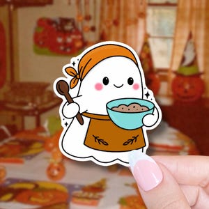 May include: A white ghost sticker with an orange headscarf and apron, holding a wooden spoon and a blue bowl filled with brown ingredients. The ghost has rosy cheeks and a smiling face. The sticker has a white border and is held by a hand.