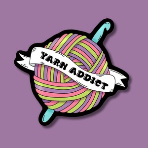 Pode incluir: Um autocolante colorido com uma bola de l&atilde; e uma agulha de croch&ecirc;. A l&atilde; &eacute; listrada com rosa, verde e roxo. Uma faixa branca que atravessa a l&atilde; diz "YARN ADDICT" em texto preto. O autocolante tem um contorno preto sobre um fundo roxo.