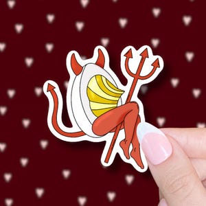 May include: A sticker featuring a cartoon devil egg character. The egg has red horns, a tail, and legs, holding a red trident. The egg's yolk is yellow and white, with a white eggshell. The background is a dark red with white hearts.