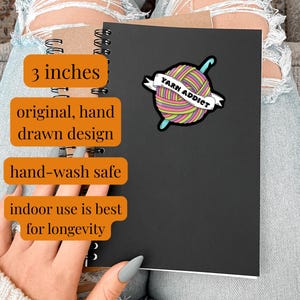 Pode incluir: Um caderno preto com um autocolante colorido "Yarn Addict". O autocolante apresenta uma bola de l&atilde; com uma agulha de croch&ecirc; e uma faixa. A imagem tamb&eacute;m inclui o texto "3 polegadas", "original, hand drawn design" e "hand-wash safe".