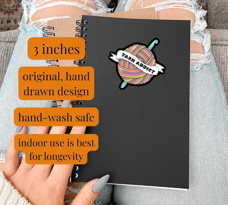 Pode incluir: Um caderno preto com um autocolante colorido "Yarn Addict". O autocolante apresenta uma bola de l&atilde; com uma agulha de croch&ecirc; e uma faixa. A imagem tamb&eacute;m inclui o texto "3 polegadas", "original, hand drawn design" e "hand-wash safe".